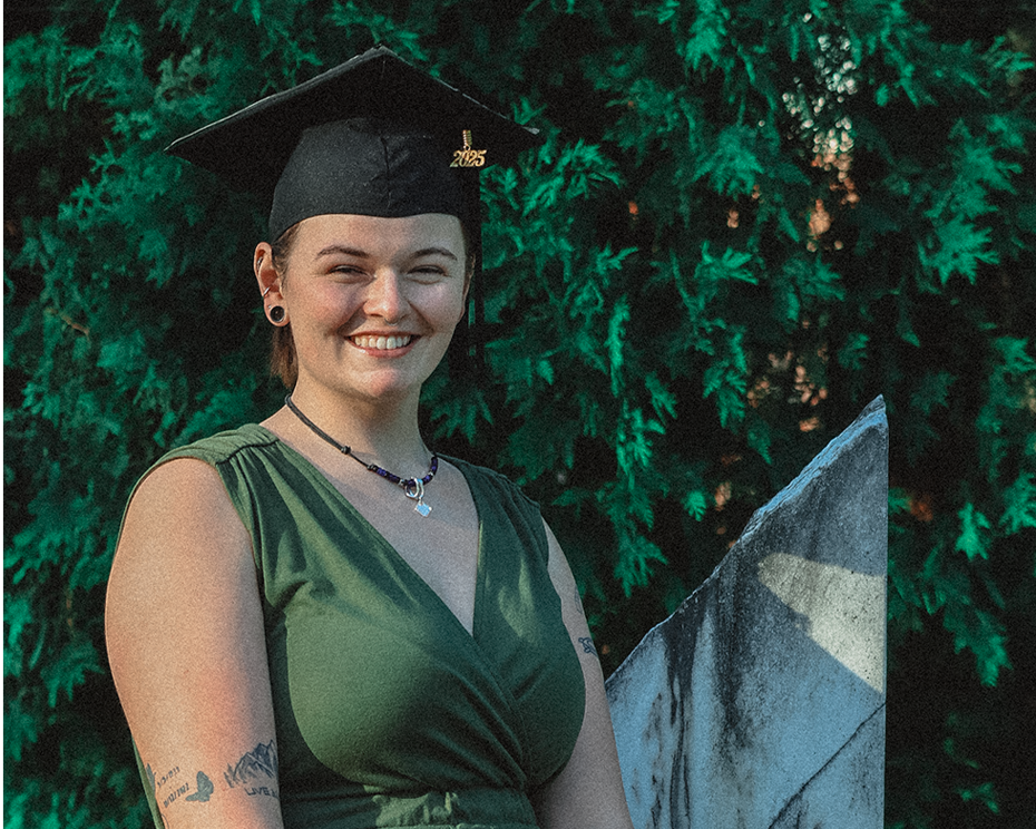A smiling young woman in a green dress and a black graduation cap with a gold '2023' emblem, sitting outdoors on a stone bench with green foliage in the background.