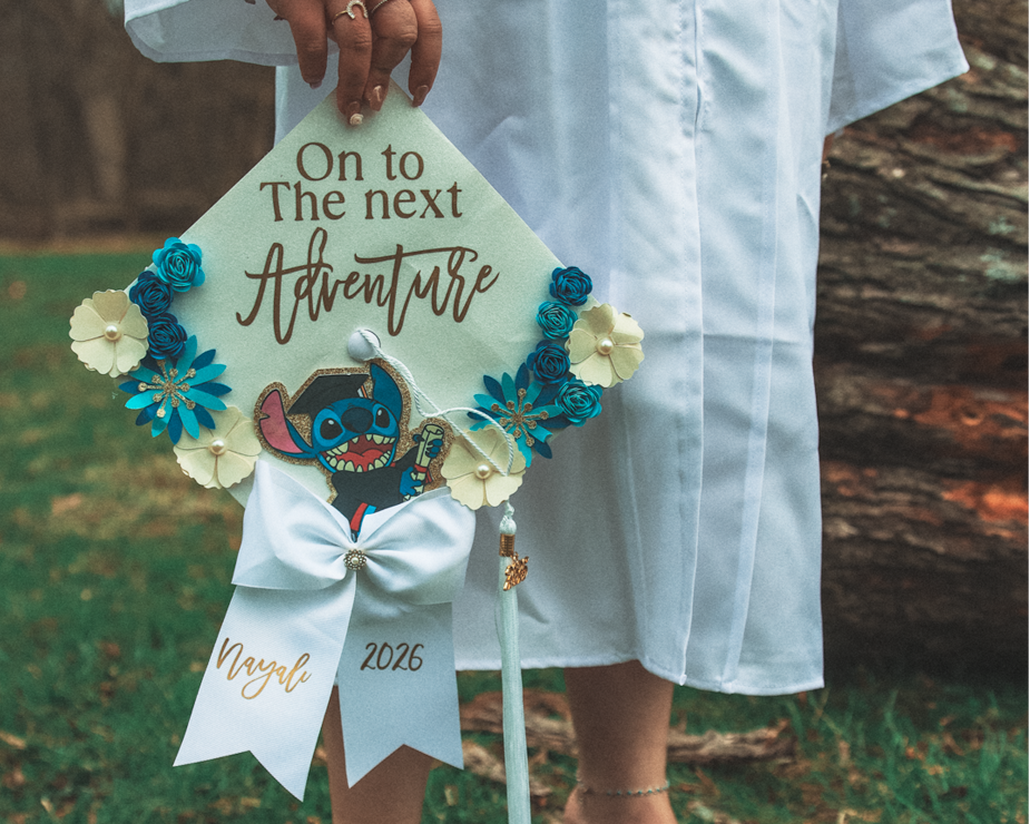 A decorated graduation cap held by a person dressed in a white gown, with a background of grass and a tree trunk. The cap is adorned with flowers, a Stitch cartoon character, and a white ribbon with gold text reading "Nayel 2026." The person is weari