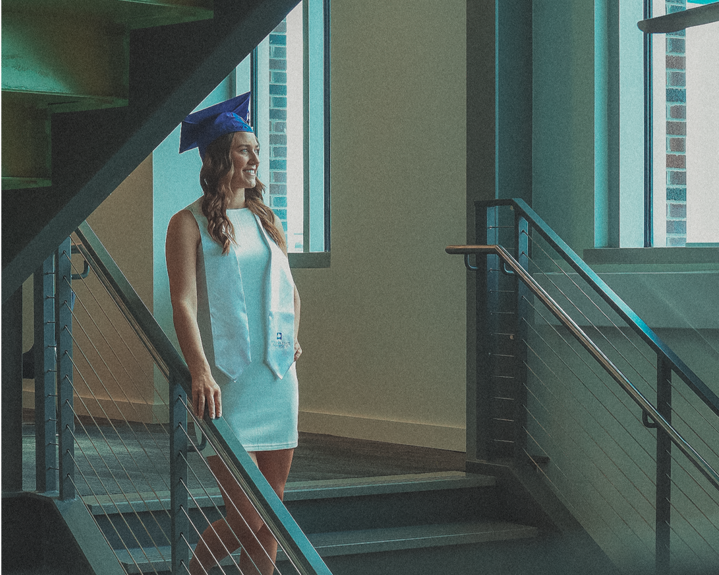 woman with a blue graduation cap and white dress standing on staircase, smiling, near large windows in a modern building.