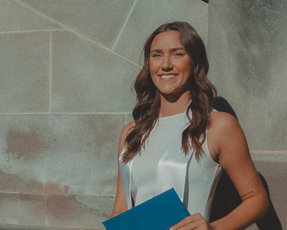 A young woman in a white dress sitting on steps outside, holding a blue diploma folder with a tassel. She is smiling with her hair down.