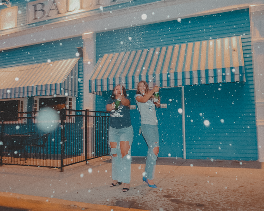 Two women standing in front of a closed storefront with blue walls and striped awnings, holding bottles and smiling while snow falls around them.
