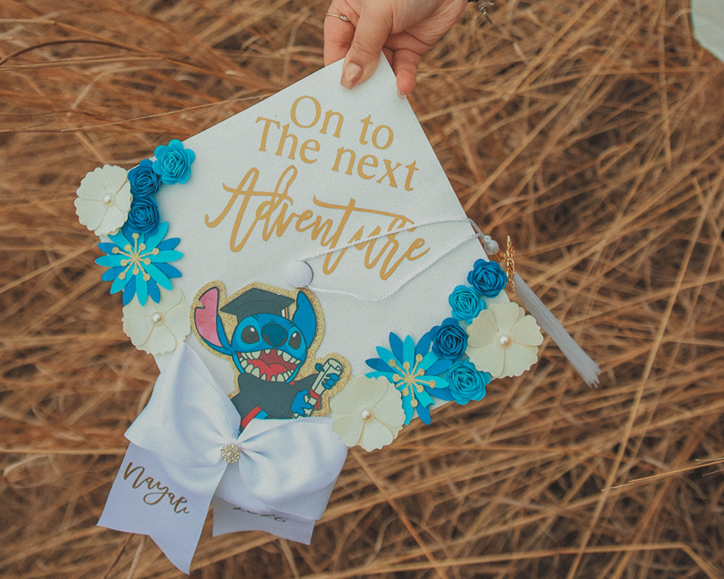A person's hand holding a decorated graduation cap with a Stitch character from Lilo & Stitch, surrounded by blue and white flowers, on a background of dry grass.