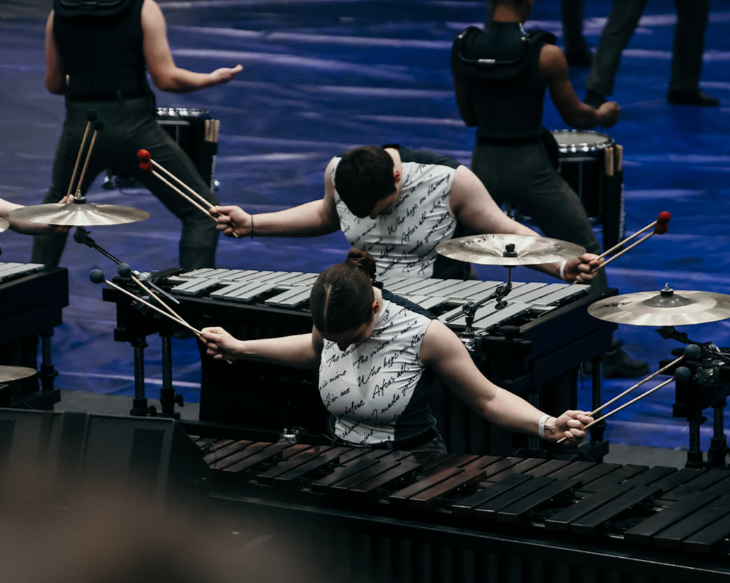 Two musicians playing percussion instruments on stage, with others in the background, illuminated by stage lighting.