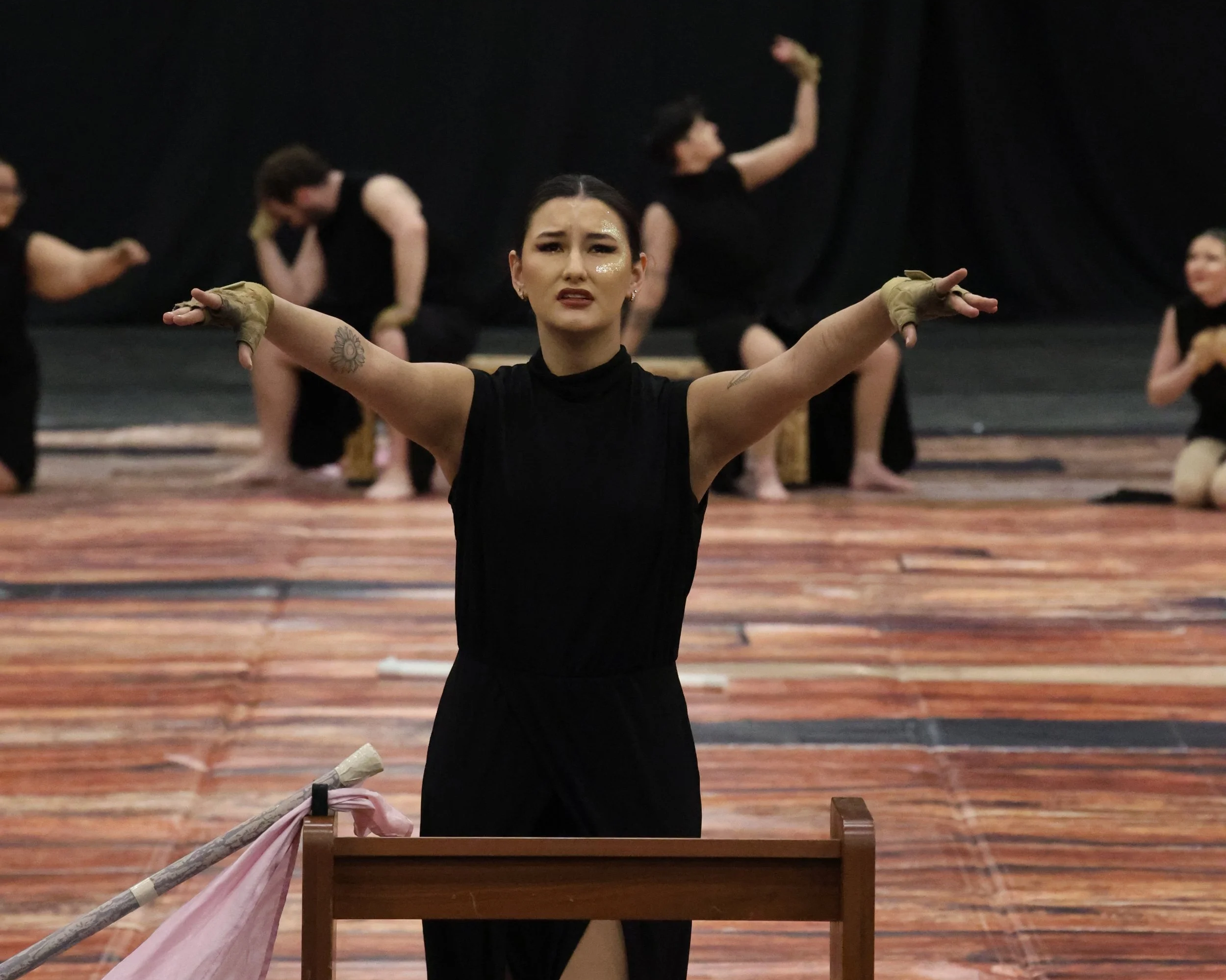A woman in black dance attire performing a dance pose with arms outstretched, in front of a black background with other women in similar attire in various poses, on a wooden stage.