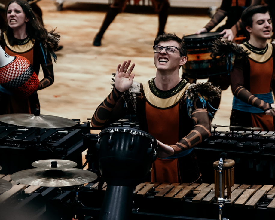 A group of musicians in costume performing with percussion instruments, including drums and xylophones, on stage. One musician is raising his hand, smiling, and wearing glasses.