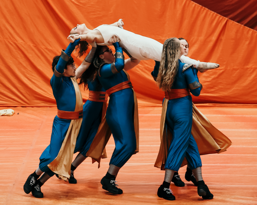 A group of performers in blue and gold costumes lift a person in a white costume during a dance or theatrical performance on a stage with an orange background.