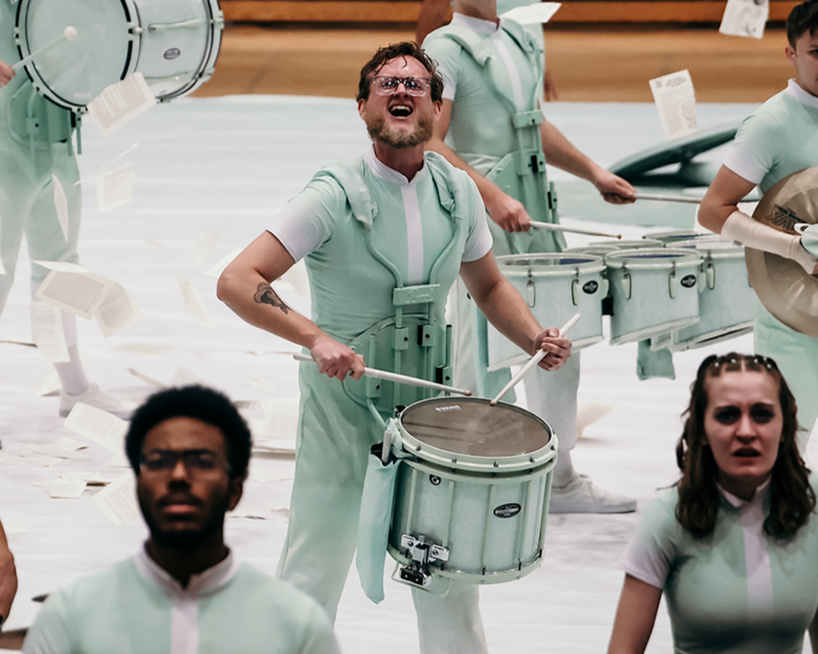 A group of musicians in light green uniforms performing with percussion instruments on a stage. Some are playing drums, and paper sheets are flying around them.