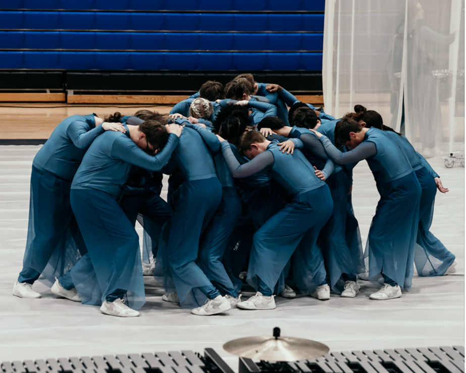 Group of figure skaters in blue costumes huddled together in a circle on the ice rink, preparing or performing a routine.