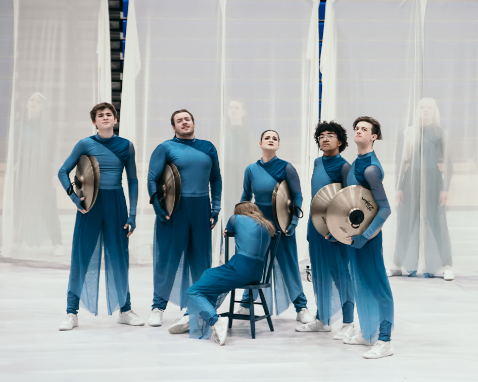 A group of six young people in matching blue outfits, holding cymbals, posing on a stage with sheer white curtains in the background.