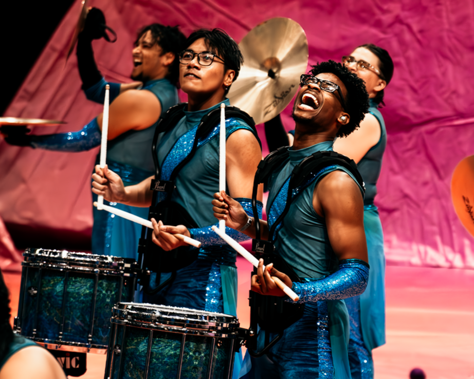 A lively group of percussionists performing on stage, dressed in matching blue and black costumes with sequined details, playing drums and cymbals.