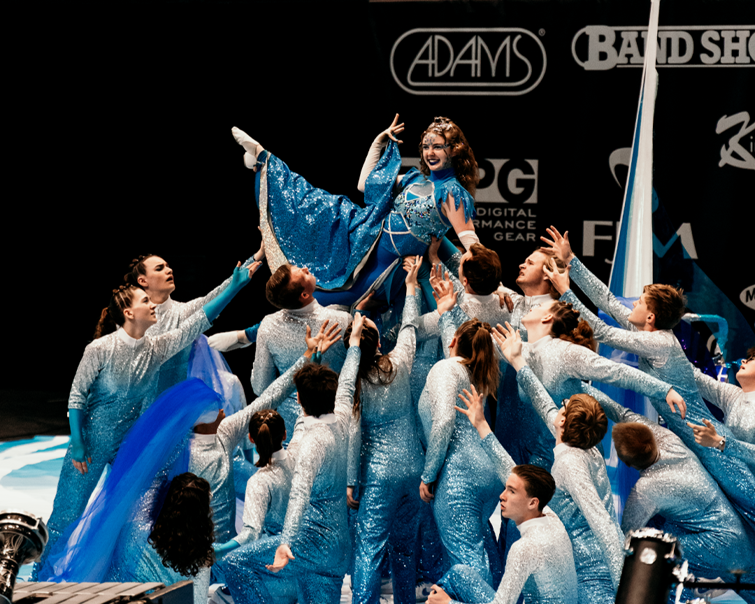 A dance team performing on stage with a woman in a blue dress being lifted by team members dressed in silver costumes.