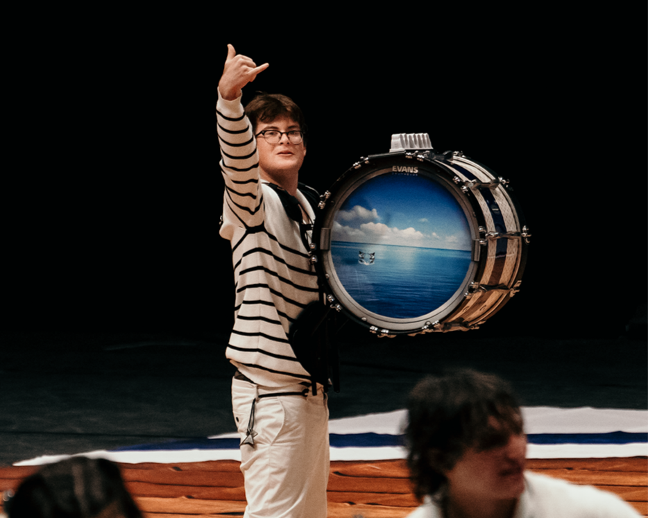 A young man holding a large drum with a painted ocean scene, standing on a wooden stage, with other people in the foreground.