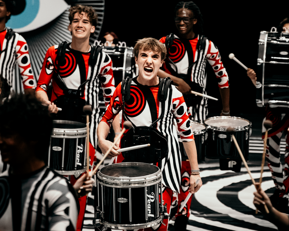 A group of young drummers performing on stage, wearing matching black, white, and red patterned outfits. The scene features a bold, black-and-white, striped floor and background decor, with a large eye design behind them.