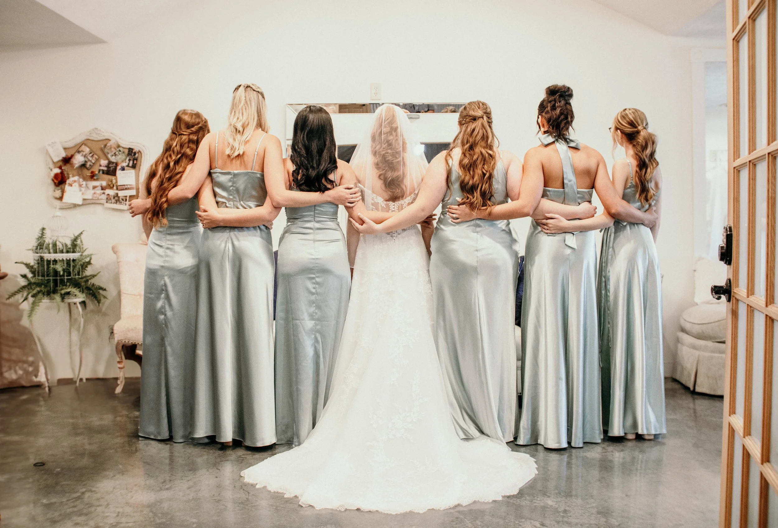 A bride and six women standing with their backs together, all wearing matching grey satin dresses, in a room with minimal decor, a small Christmas tree to the left, and a bulletin board with photos on the wall.