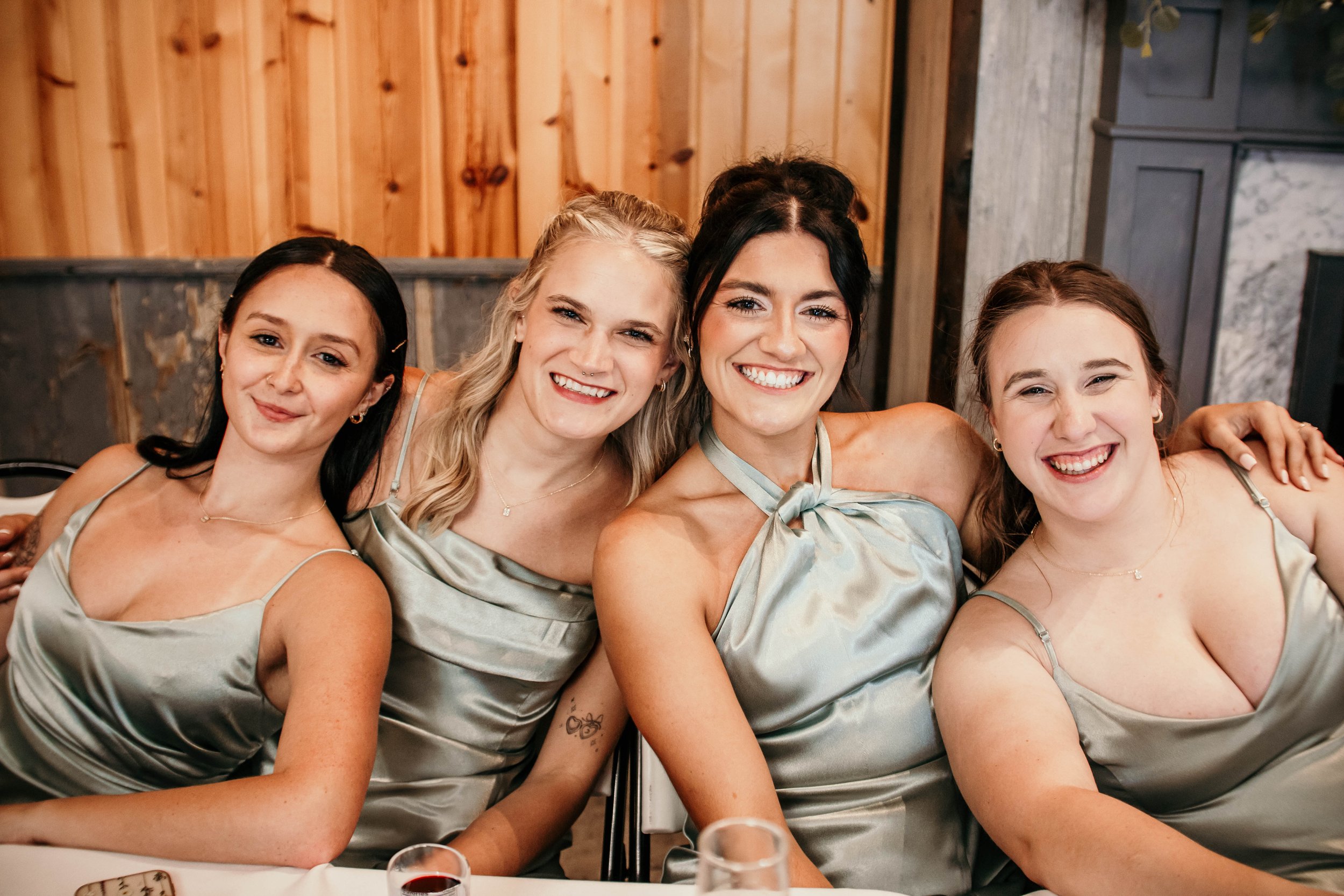 Four women in satin dresses sitting closely together at a dinner table, smiling for the camera. They are indoors, with a wooden wall in the background and drinks on the table.