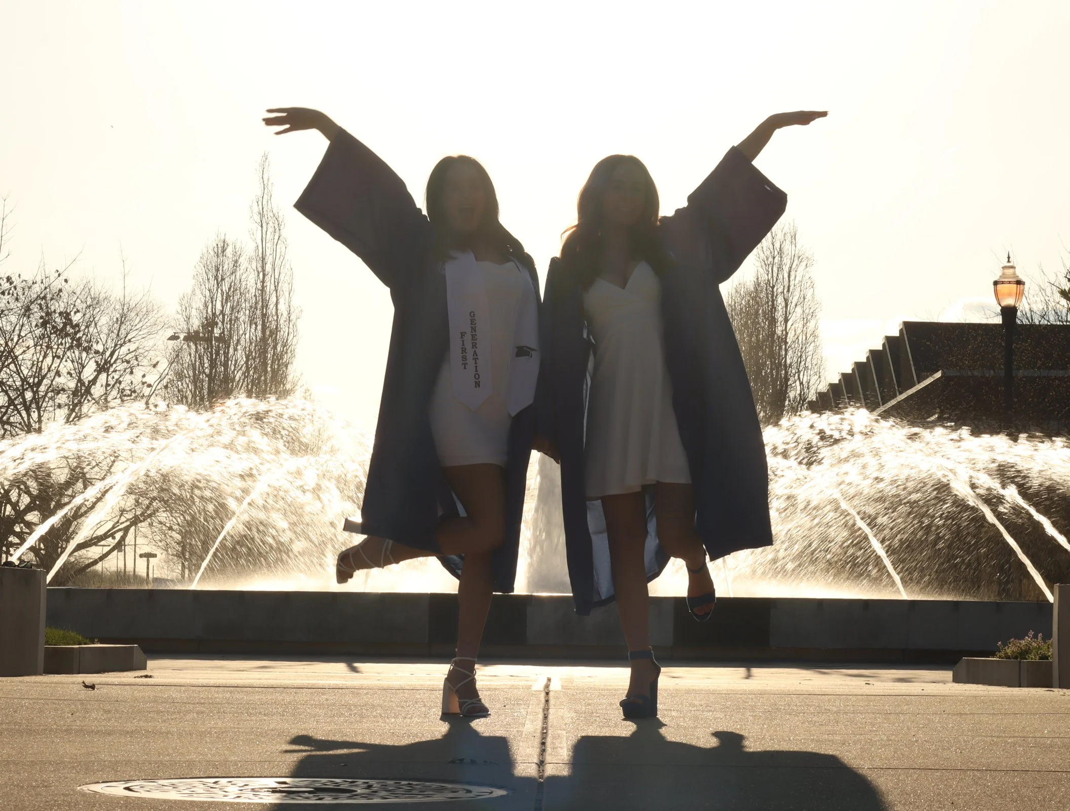 Two women in graduation gowns and caps stand in front of a fountain during sunset, smiling and posing with arms outstretched.