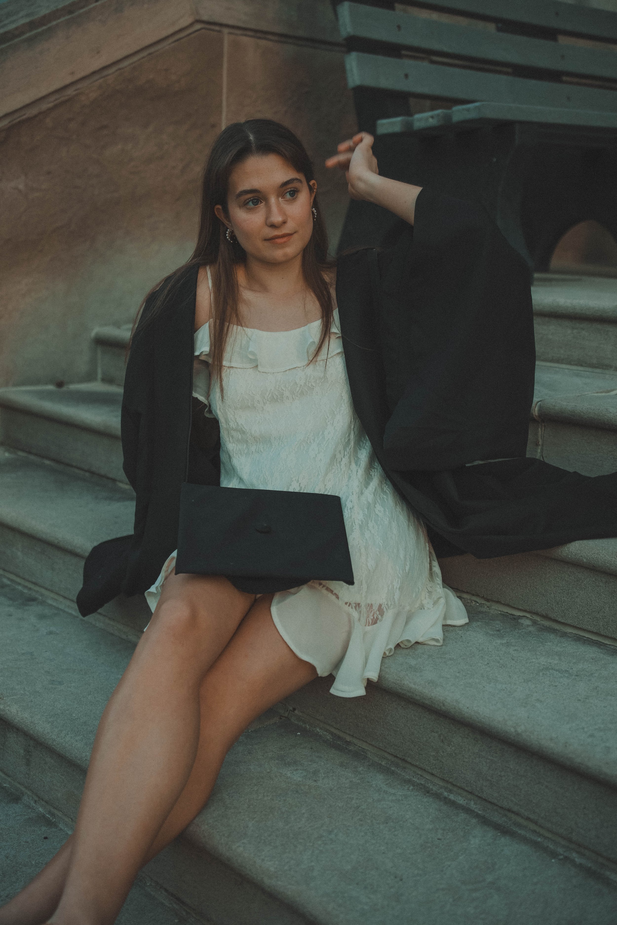 A woman sitting on stone steps outdoors, holding a black purse and fully dressed in a white dress, with her left hand resting on her head.