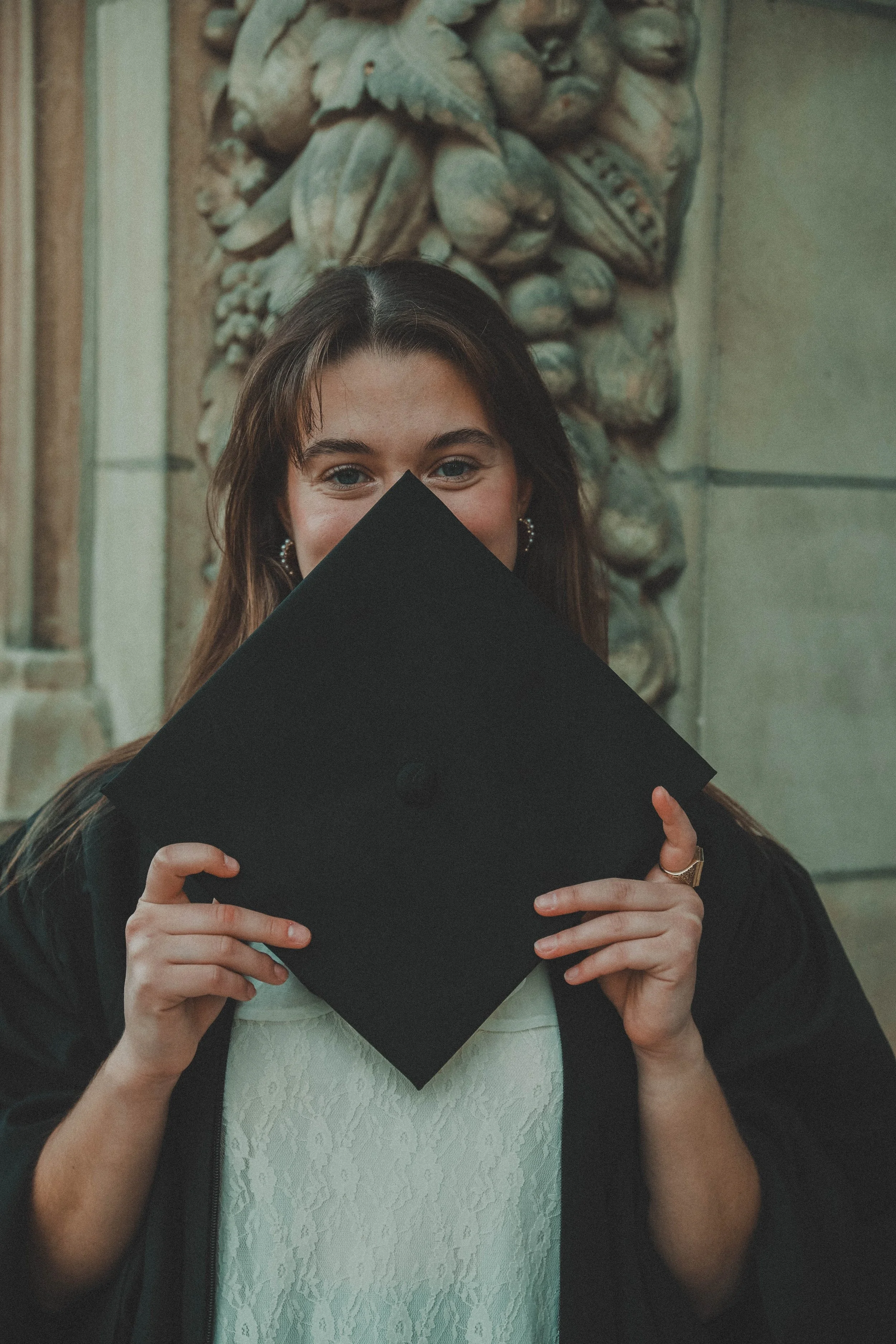 A young woman holding a black graduation cap in front of her face, with only her eyes and part of her forehead visible, standing in front of a textured stone wall.