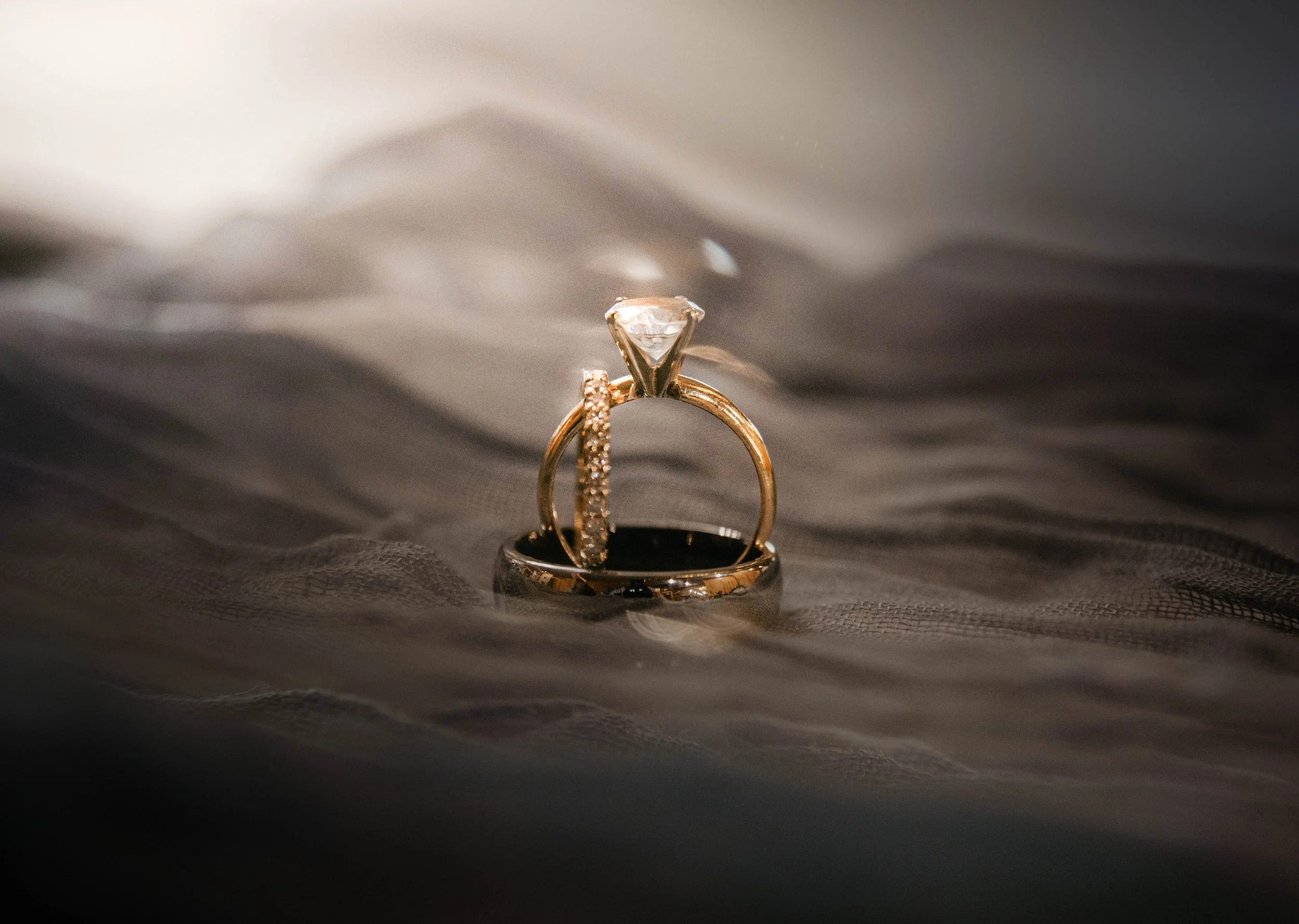 A close-up of a diamond engagement ring with a solitaire stone, surrounded by a diamond-studded band, resting on a dark fabric surface.