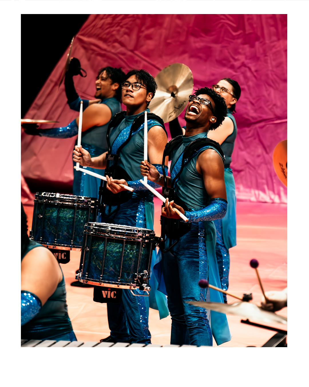A lively group of percussionists performing on stage, dressed in matching blue and black costumes with sequined details, playing drums and cymbals.