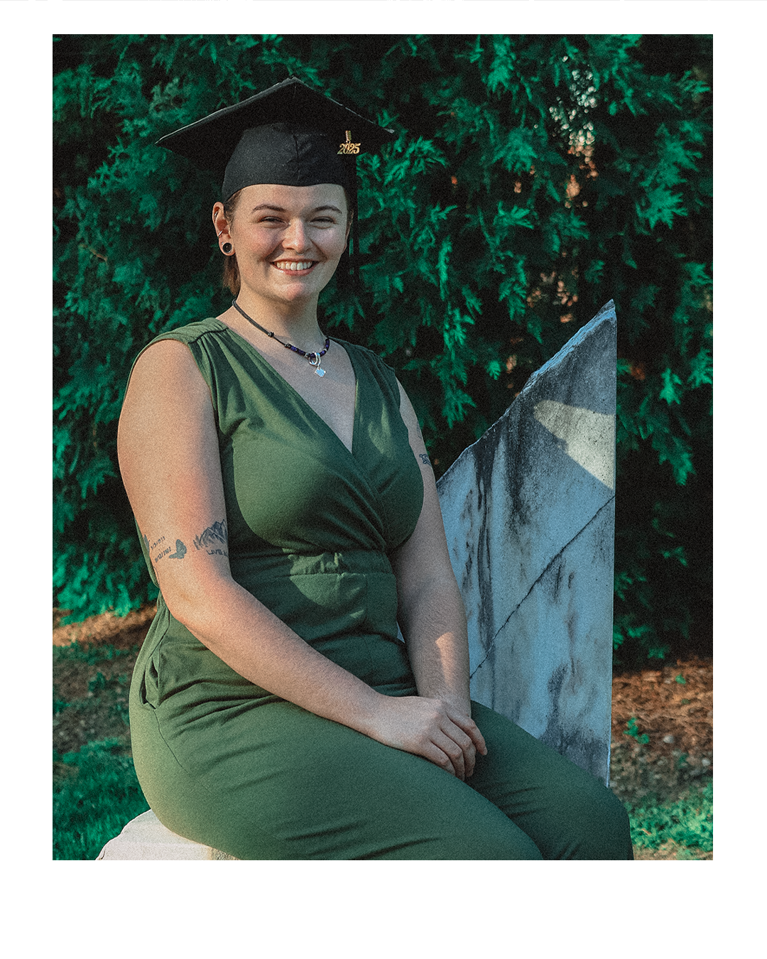A smiling young woman in a green dress and a black graduation cap with a gold '2023' emblem, sitting outdoors on a stone bench with green foliage in the background.