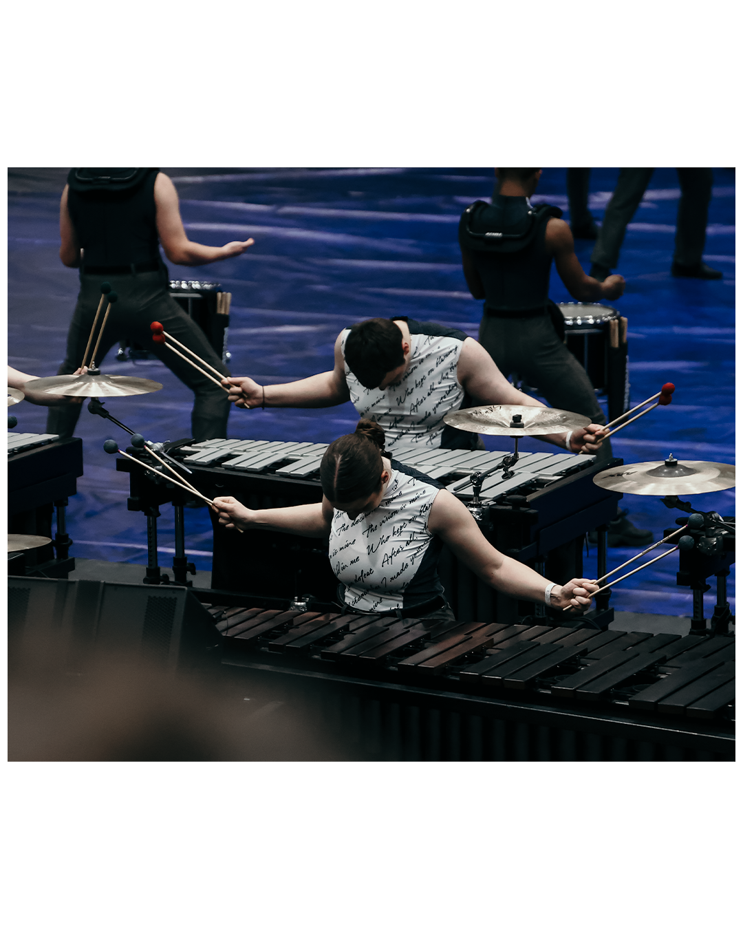 Two musicians playing percussion instruments on stage, with others in the background, illuminated by stage lighting.