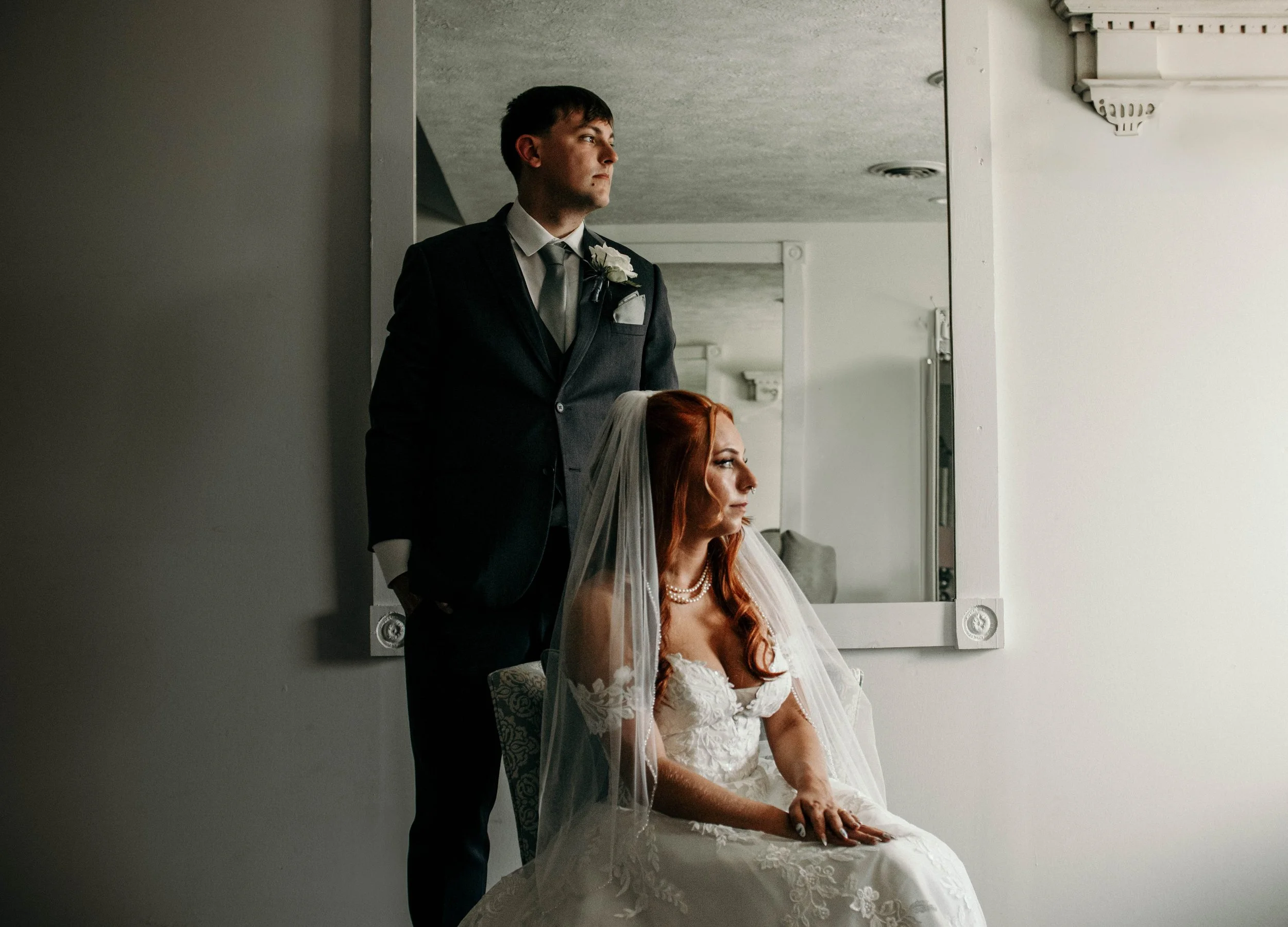 A groom in a black suit with a white boutonniere looks out a window while a bride with red hair, wearing a white lace wedding dress and pearl necklace, sits nearby.