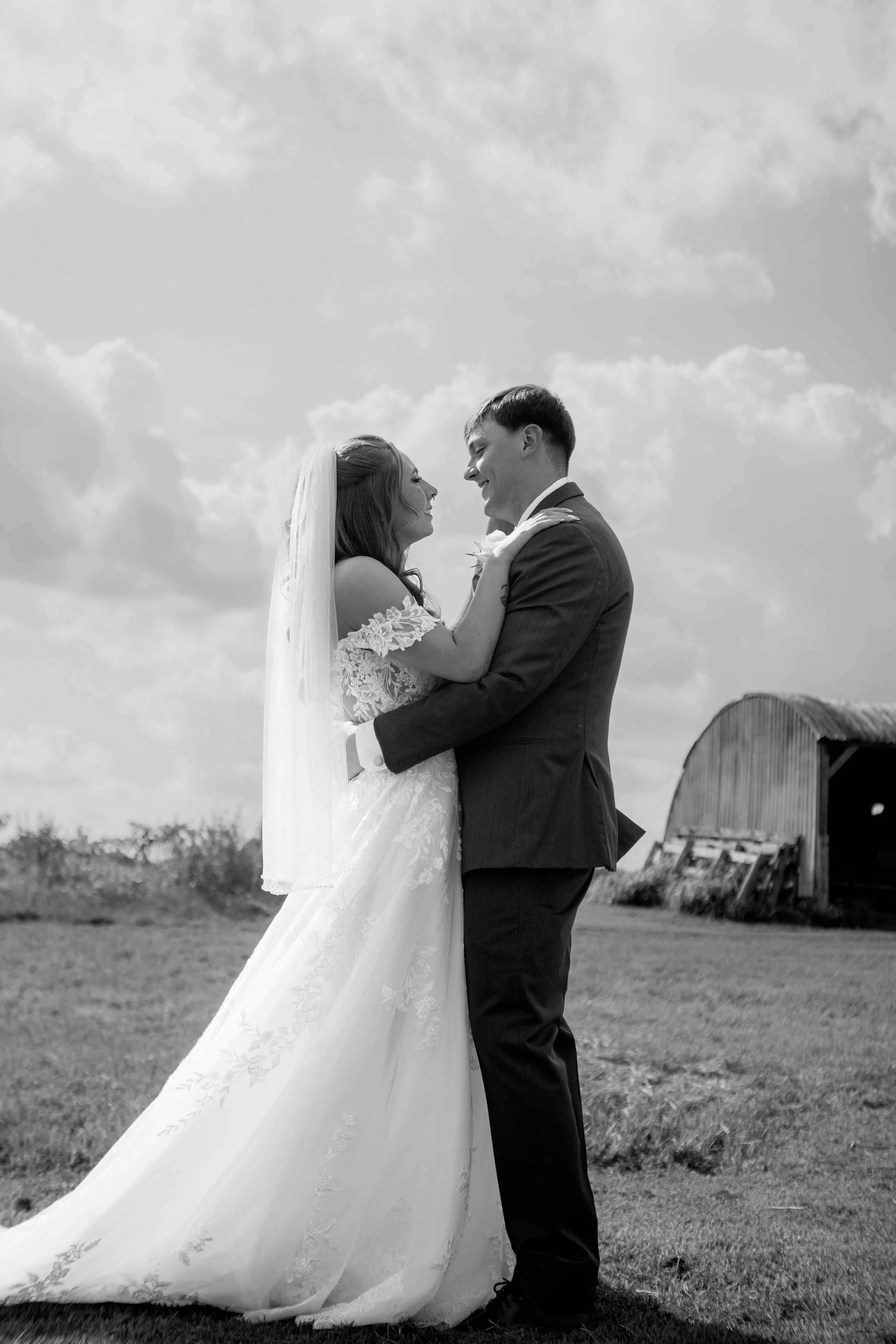 A black and white photo of a newlywed couple outside in a rural area with a barn in the background. The bride is wearing a wedding gown and veil, and the groom is in a suit. They are facing each other, smiling and holding each other tenderly.