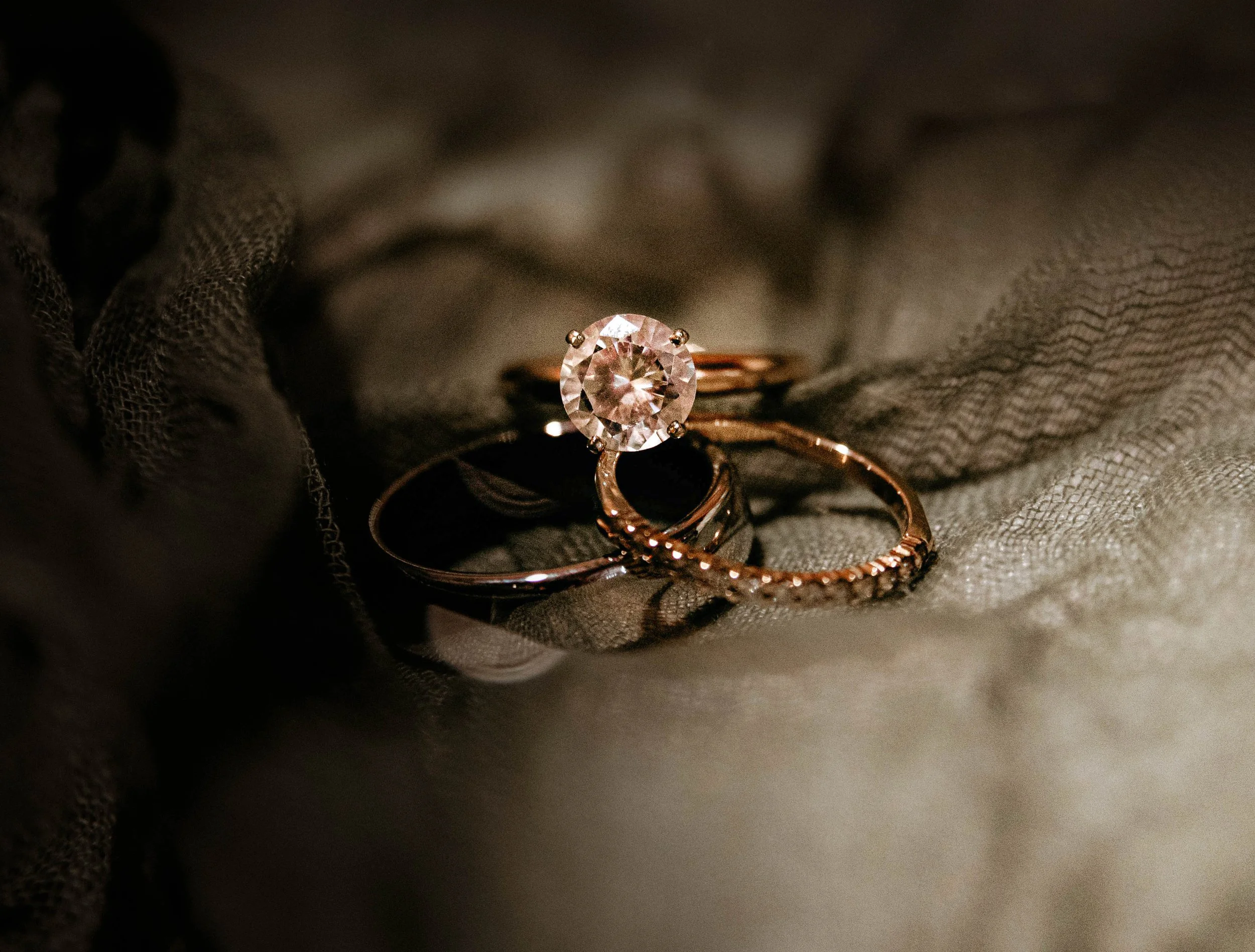 A close-up of a diamond engagement ring with a large round center stone, set in a prong setting, with a rose gold band, laid on soft, textured fabric.