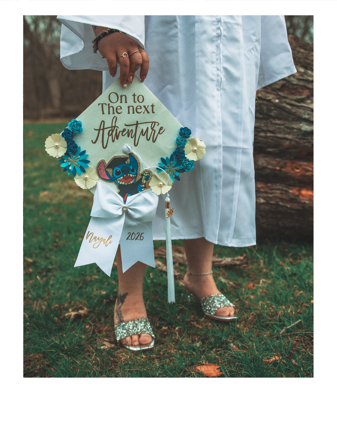 A decorated graduation cap held by a person dressed in a white gown, with a background of grass and a tree trunk. The cap is adorned with flowers, a Stitch cartoon character, and a white ribbon with gold text reading "Nayel 2026." The person is weari