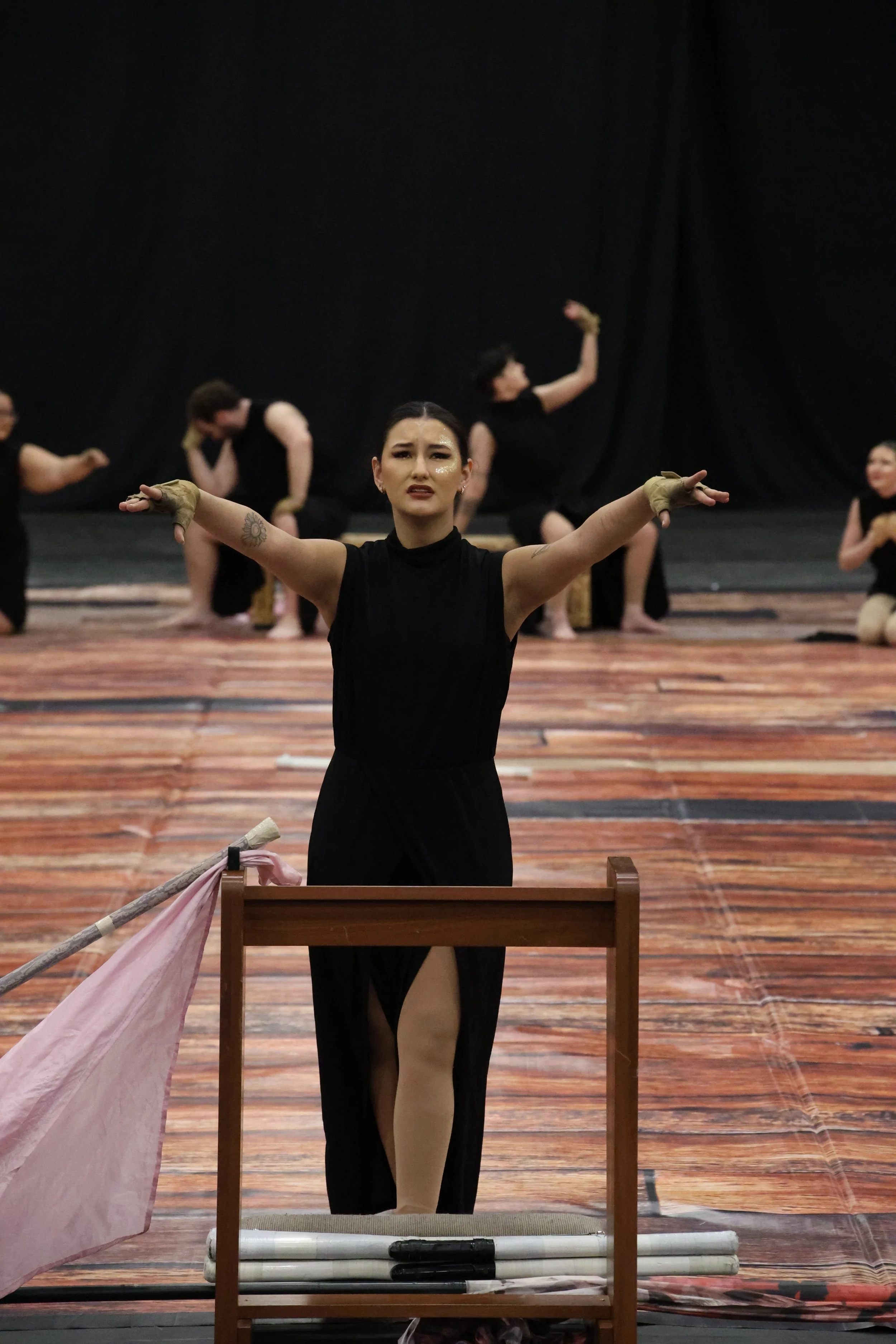 A woman in black dance attire performing a dance pose with arms outstretched, in front of a black background with other women in similar attire in various poses, on a wooden stage.