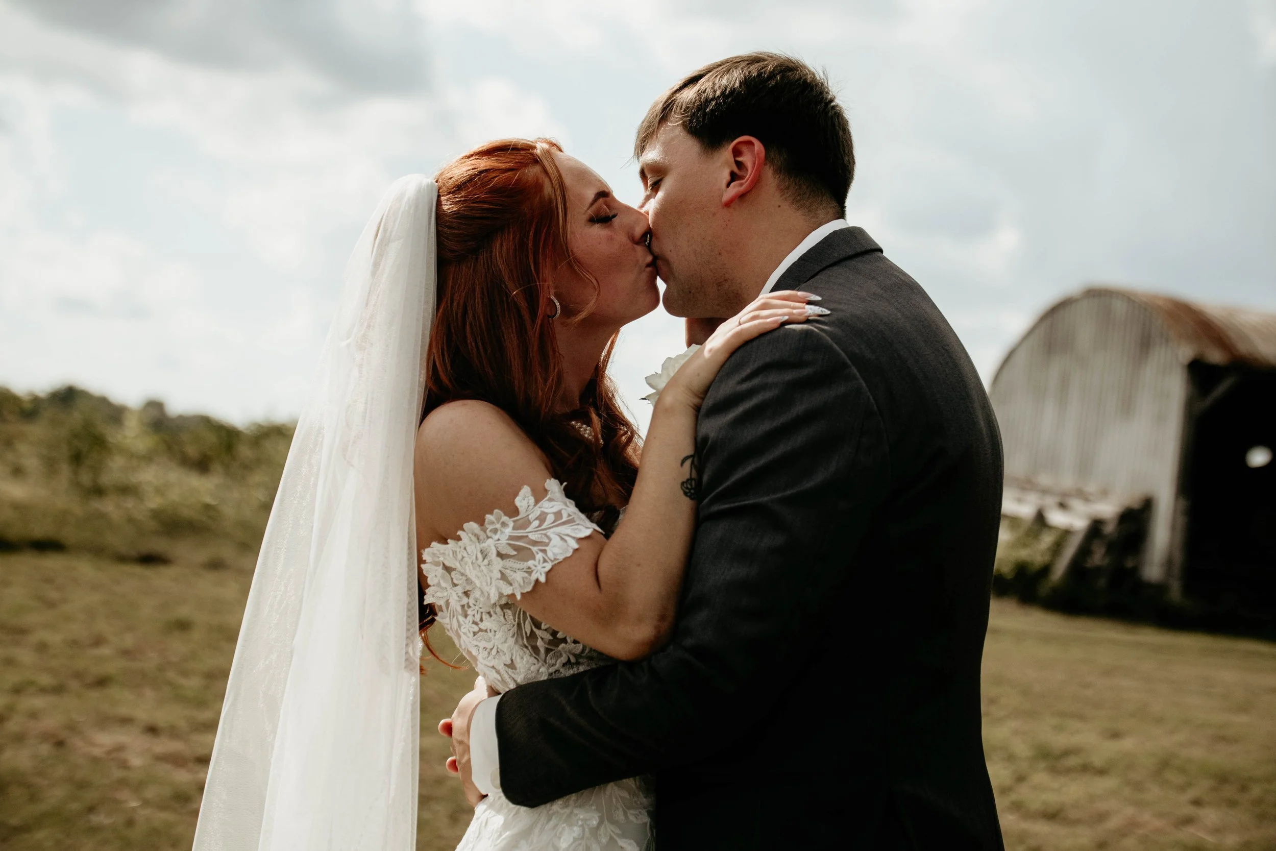 A couple is kissing outdoors, with the woman in a lace wedding dress and veil and the man in a dark suit, during daytime under cloudy sky.