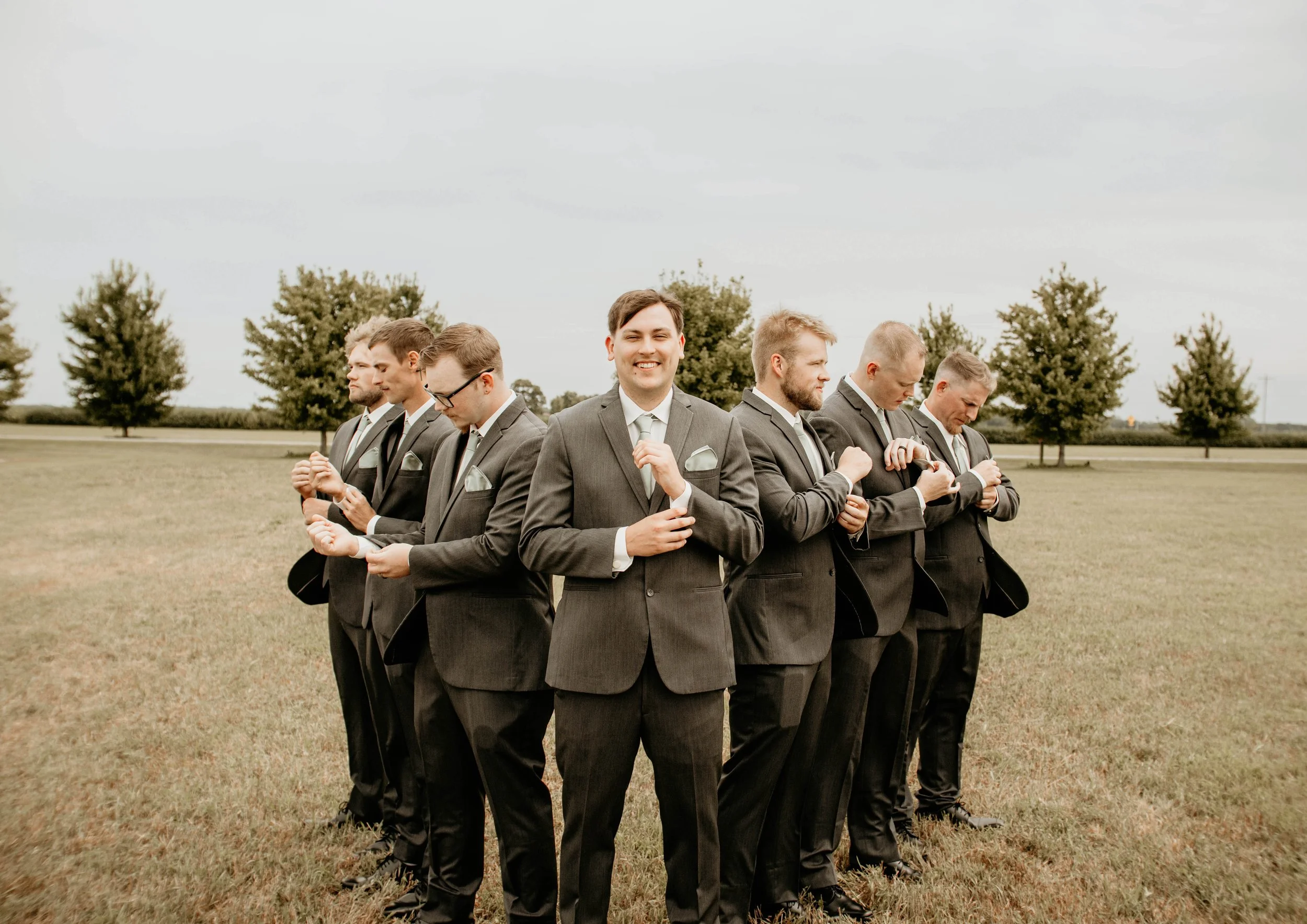 A group of men in suits standing in a line outdoors on a grassy field with trees in the background, some looking down or to the side, and one smiling at the camera.