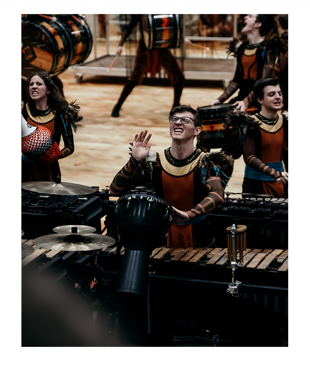 A group of musicians in costume performing with percussion instruments, including drums and xylophones, on stage. One musician is raising his hand, smiling, and wearing glasses.