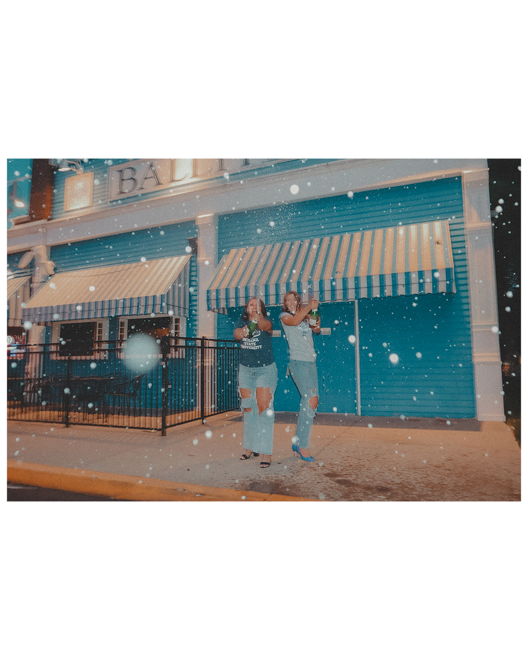 Two women standing in front of a closed storefront with blue walls and striped awnings, holding bottles and smiling while snow falls around them.