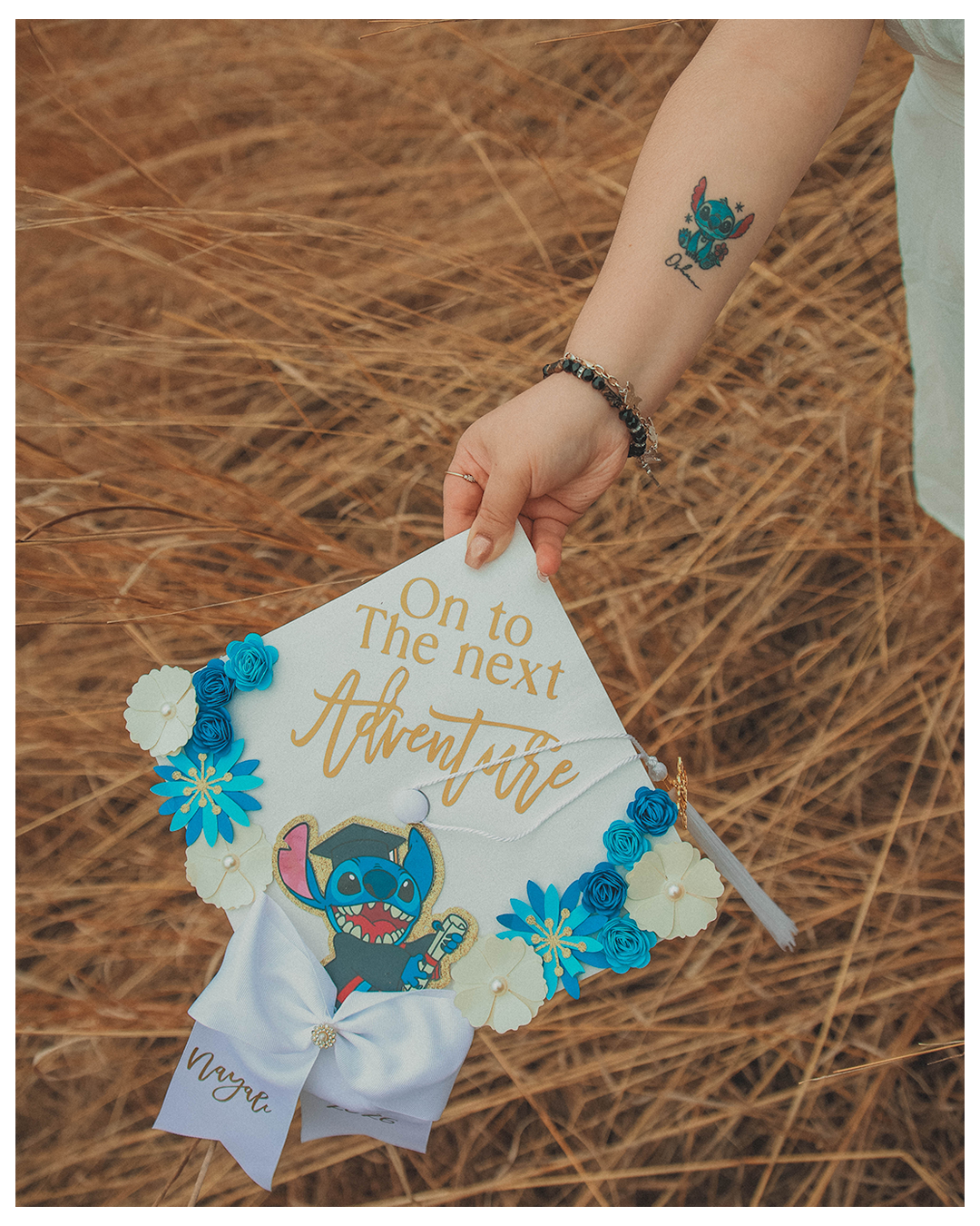A person's hand holding a decorated graduation cap with a Stitch character from Lilo & Stitch, surrounded by blue and white flowers, on a background of dry grass.