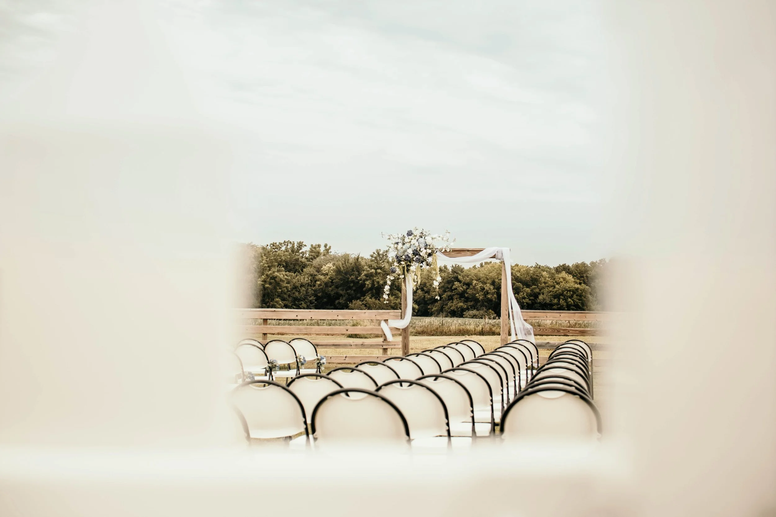 An outdoor wedding setup with chairs arranged facing a wooden arch decorated with white fabric and flowers, set in a natural environment with trees under a cloudy sky.