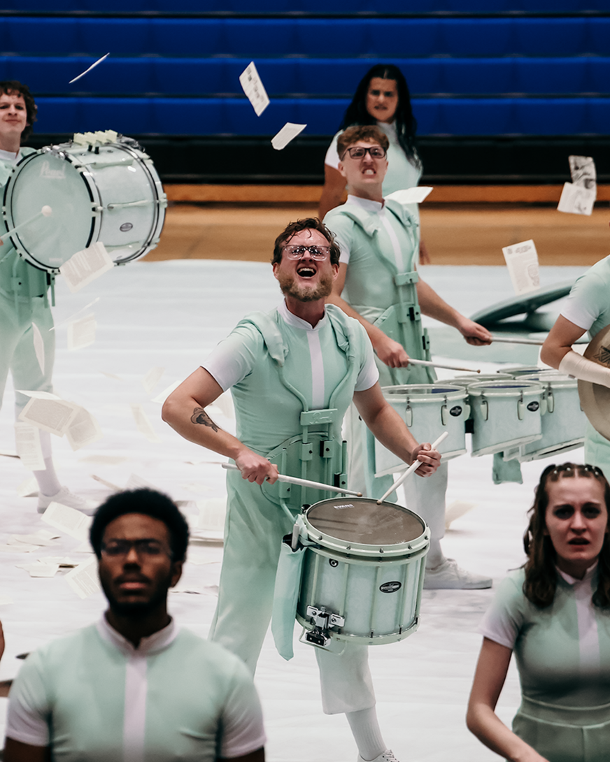 A group of drummers playing drums during a performance in a gymnasium. They are dressed in matching light green outfits and appear energetic, with some smiling and others showing intense focus. Books and papers are flying around them.