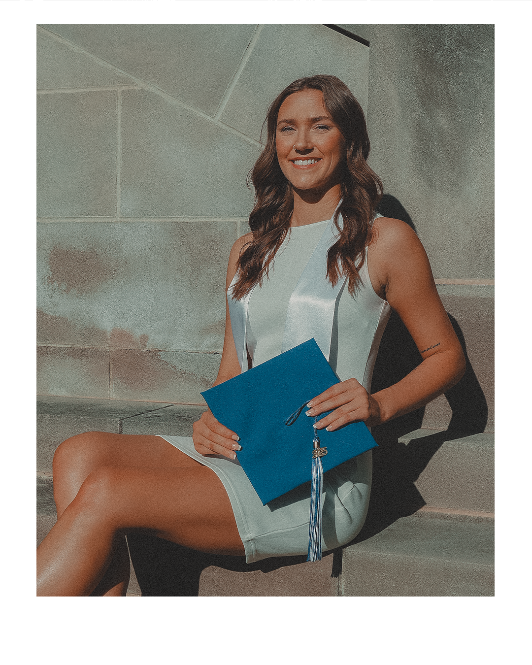 A young woman in a white dress sitting on steps outside, holding a blue diploma folder with a tassel. She is smiling with her hair down.