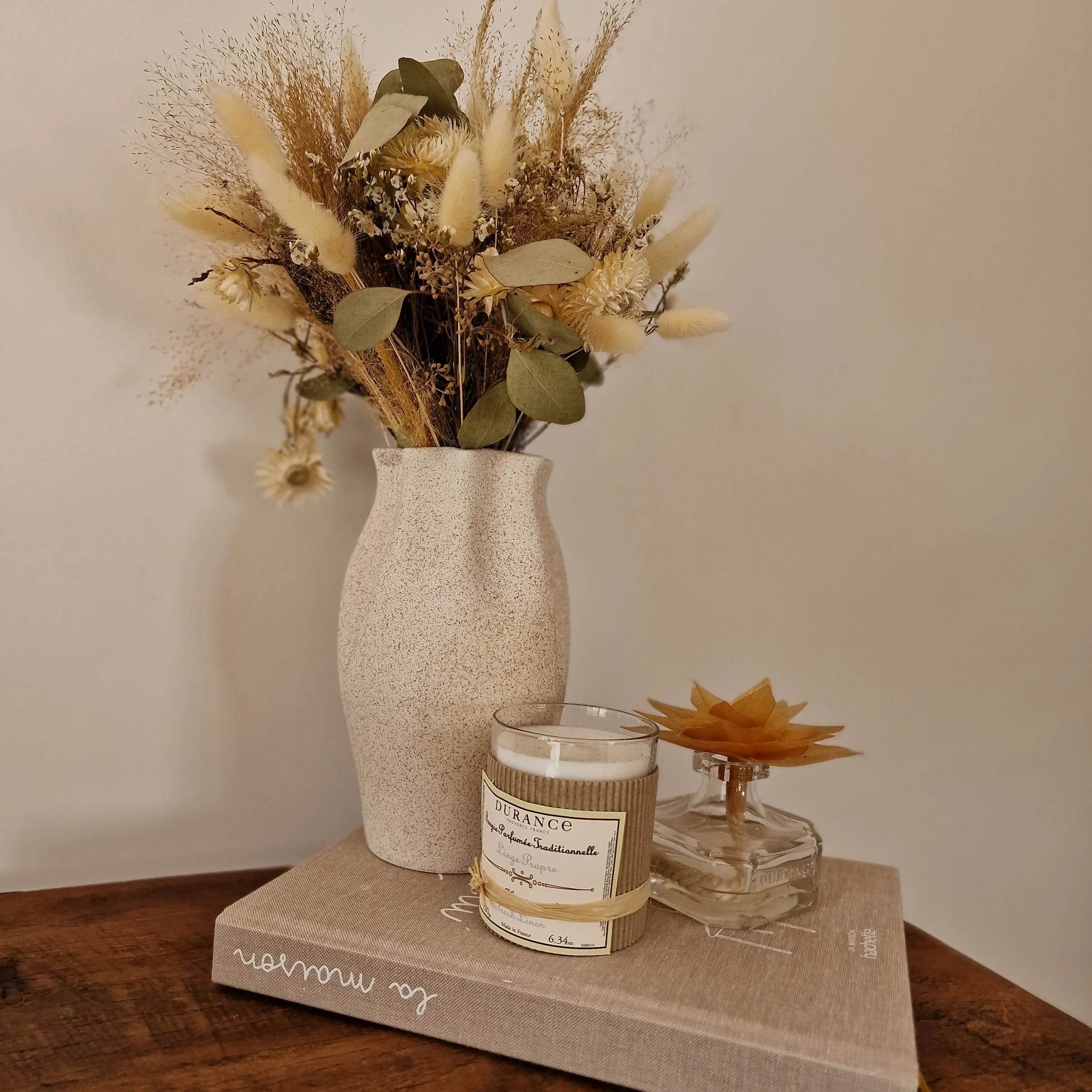 Un bouquet de fleurs séchées dans un vase blanc et un pot de miel sont placés sur une table en bois.