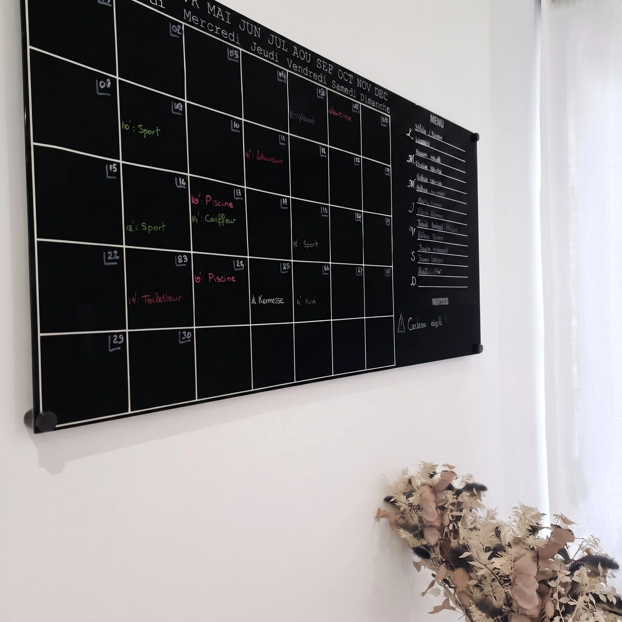 Un tableau noir d'agenda mural avec des jours et des activités écrites, situé dans une pièce lumineuse avec une fenêtre à droite. Une plante décorative se trouve en bas à droite.