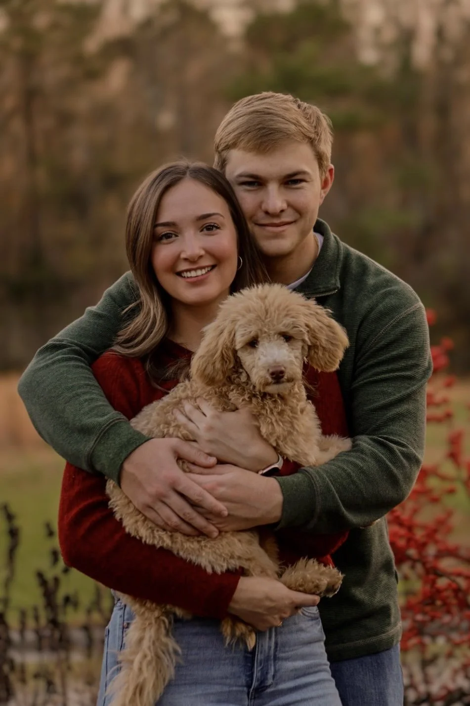 A young couple smiling and holding a fluffy apricot-colored poodle outdoors during autumn, with trees and red leaves in the background.