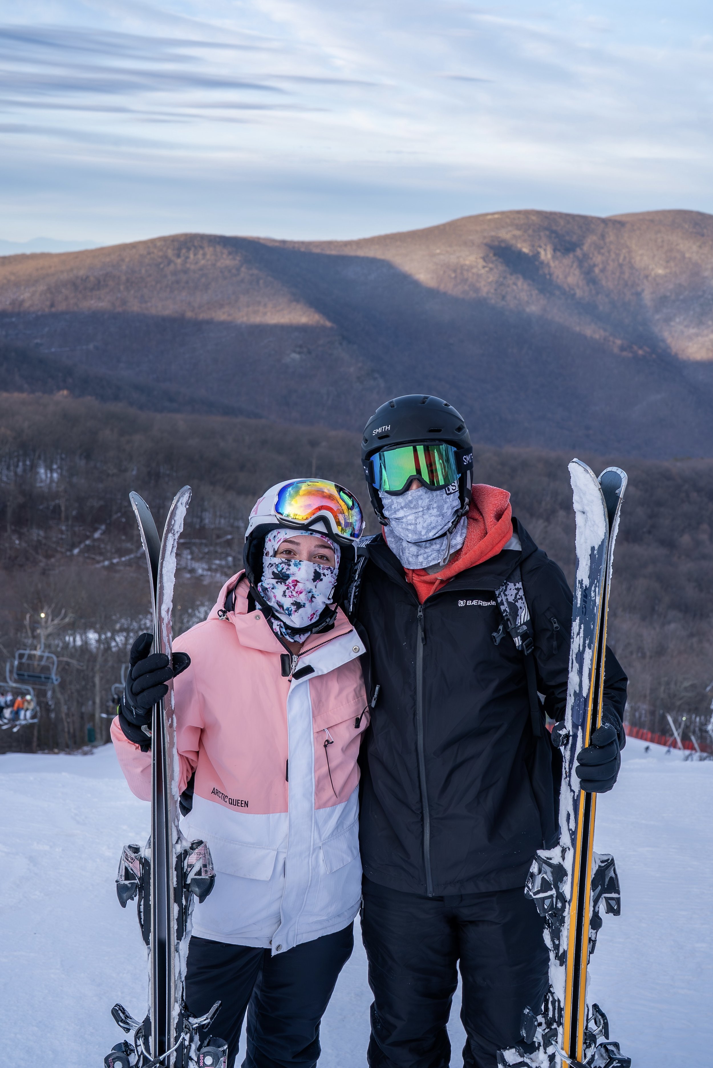 A couple in ski gear standing on a snow-covered slope with mountains in the background during daytime.