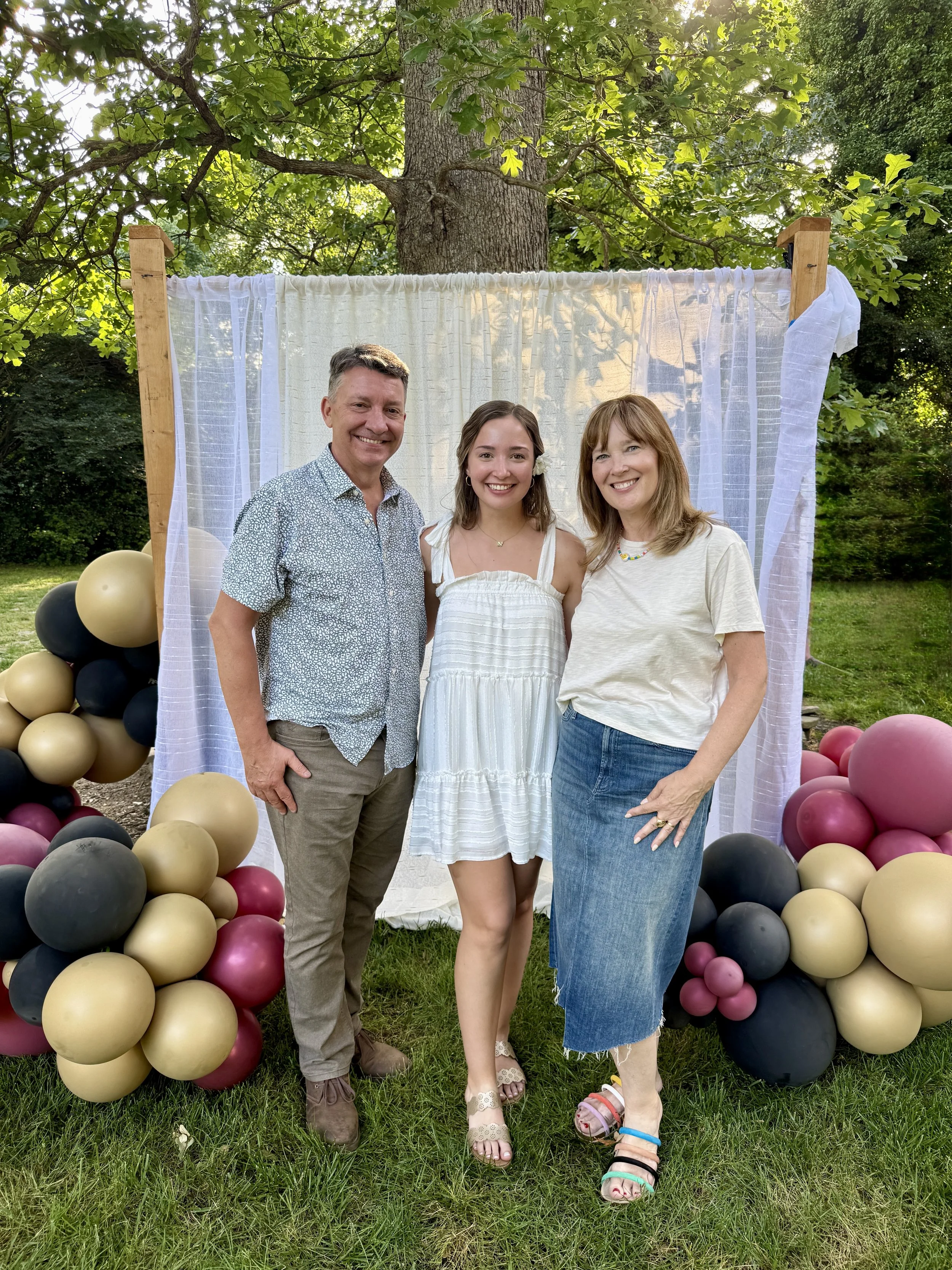 Three smiling people standing outdoors in front of a decorated backdrop with balloons on each side.