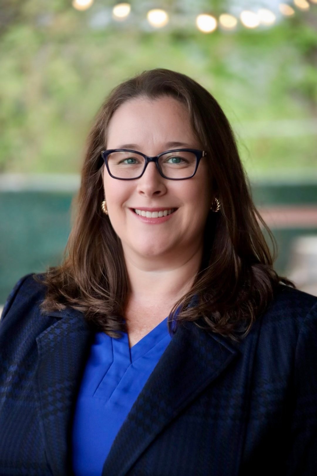 Portrait of a woman with glasses, brown hair, and earrings, smiling, wearing a blue top and dark blazer, with a blurred outdoor background.