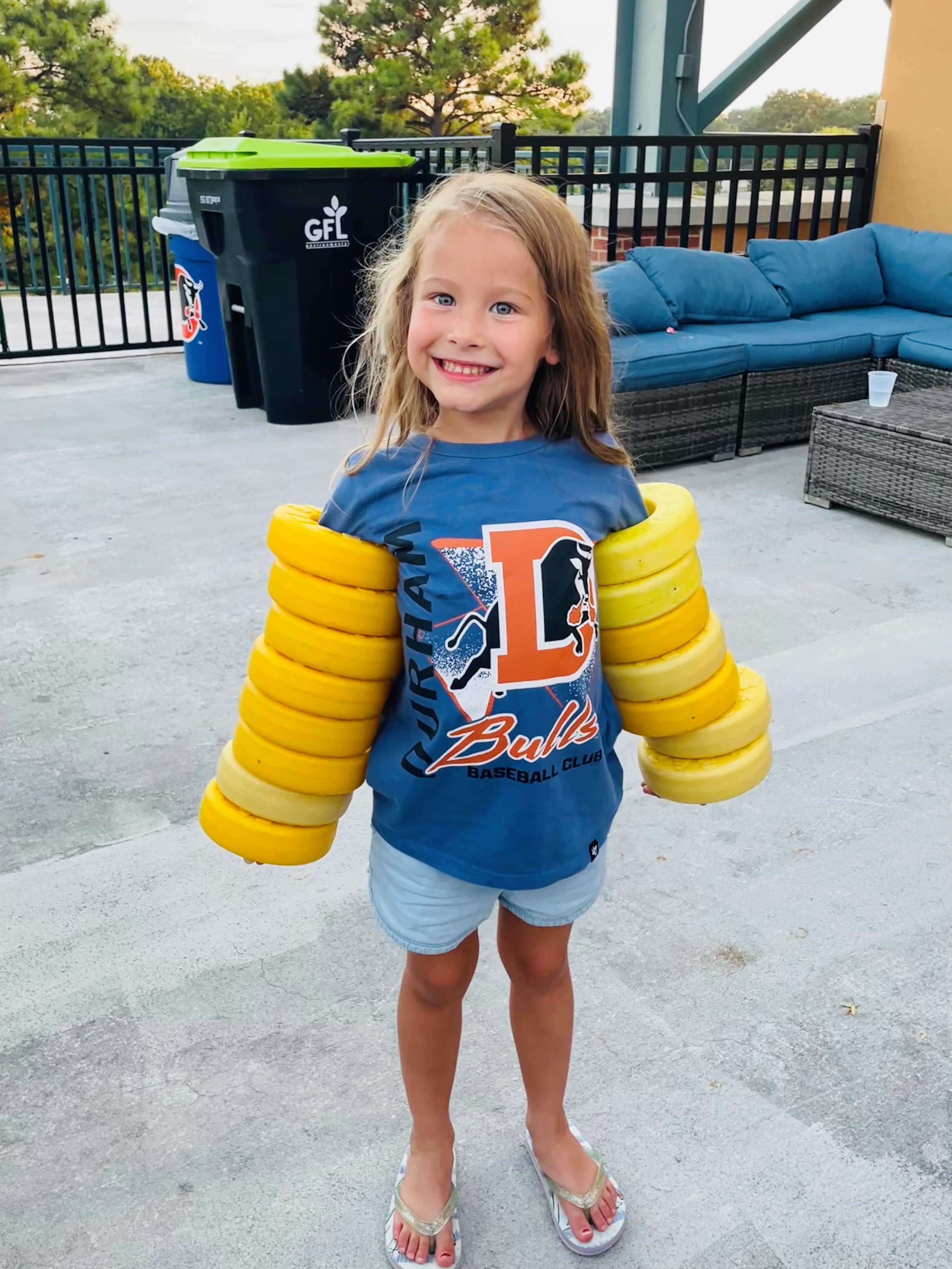 A young girl with long blonde hair, smiling, wearing a blue T-shirt with a sports team logo and shorts, standing on an outdoor patio with a wicker sofa and a cooler in the background, holding yellow frisbee discs on her arms.