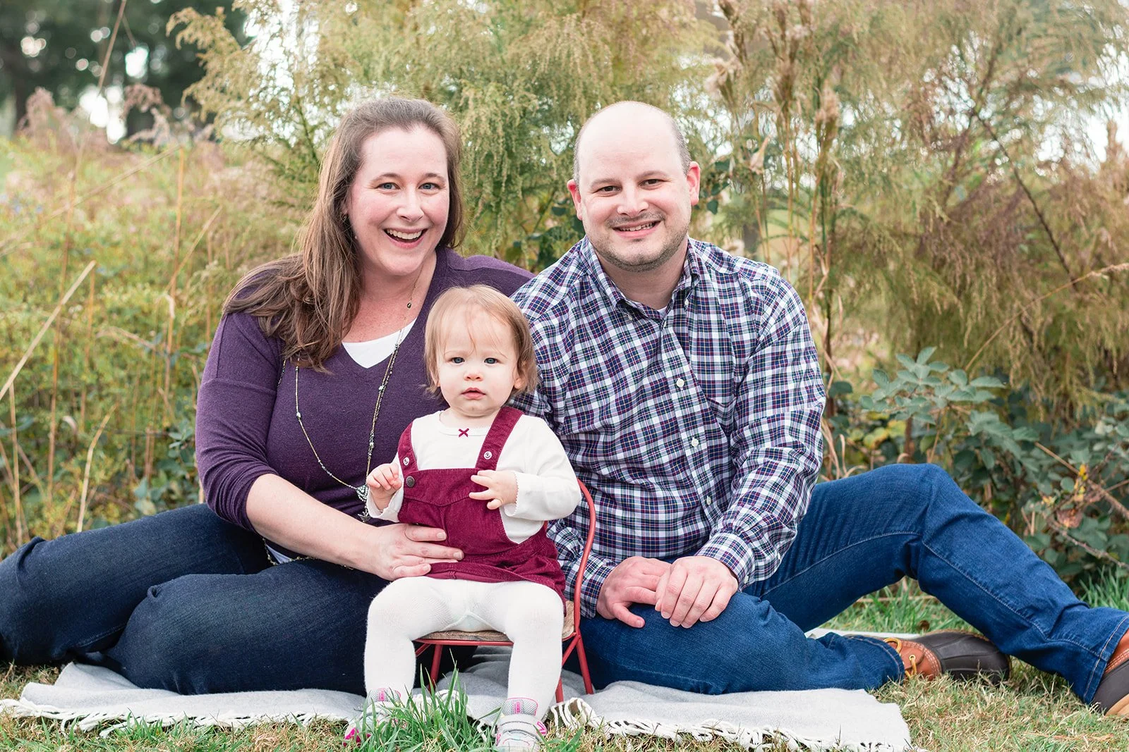 A smiling family of three sitting on a blanket outdoors during fall. The woman has long brown hair, the man has a bald head, and their young daughter has light brown hair and is wearing a white top with maroon overalls. They are surrounded by green and yellow foliage.