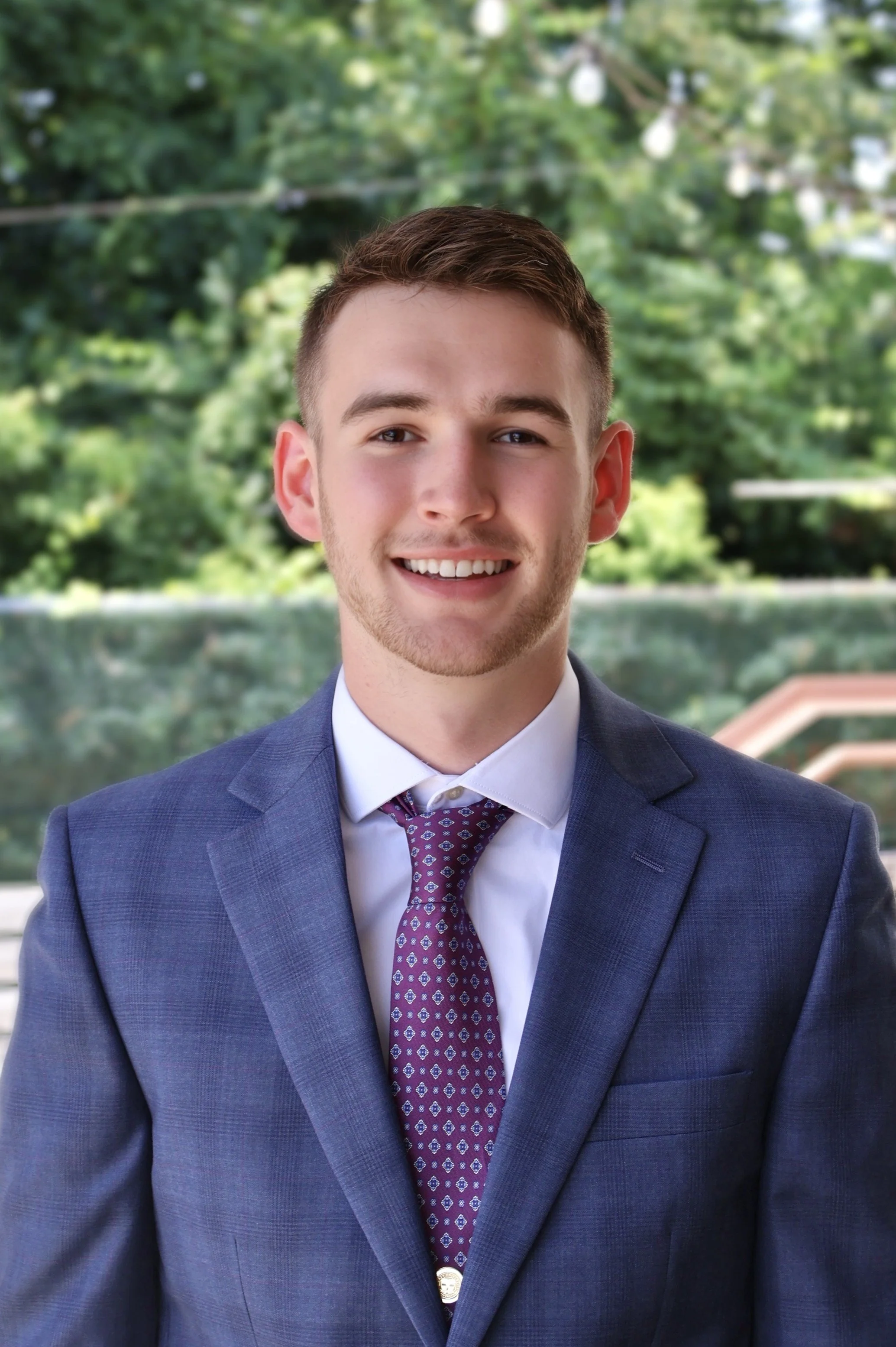 A young man with short brown hair and a light beard, wearing a blue suit, white shirt, and purple patterned tie, smiling outdoors with green trees in the background.
