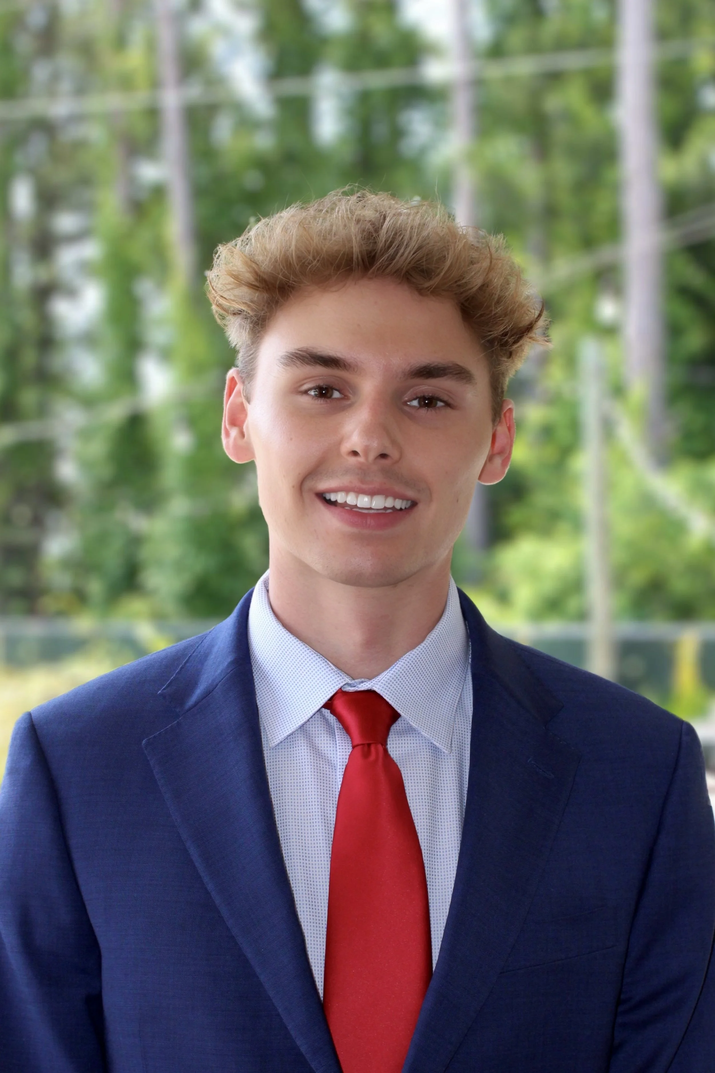 A young man with blonde hair wearing a navy blue suit, light blue shirt, and red tie, smiling outdoors with a background of green trees and power lines.