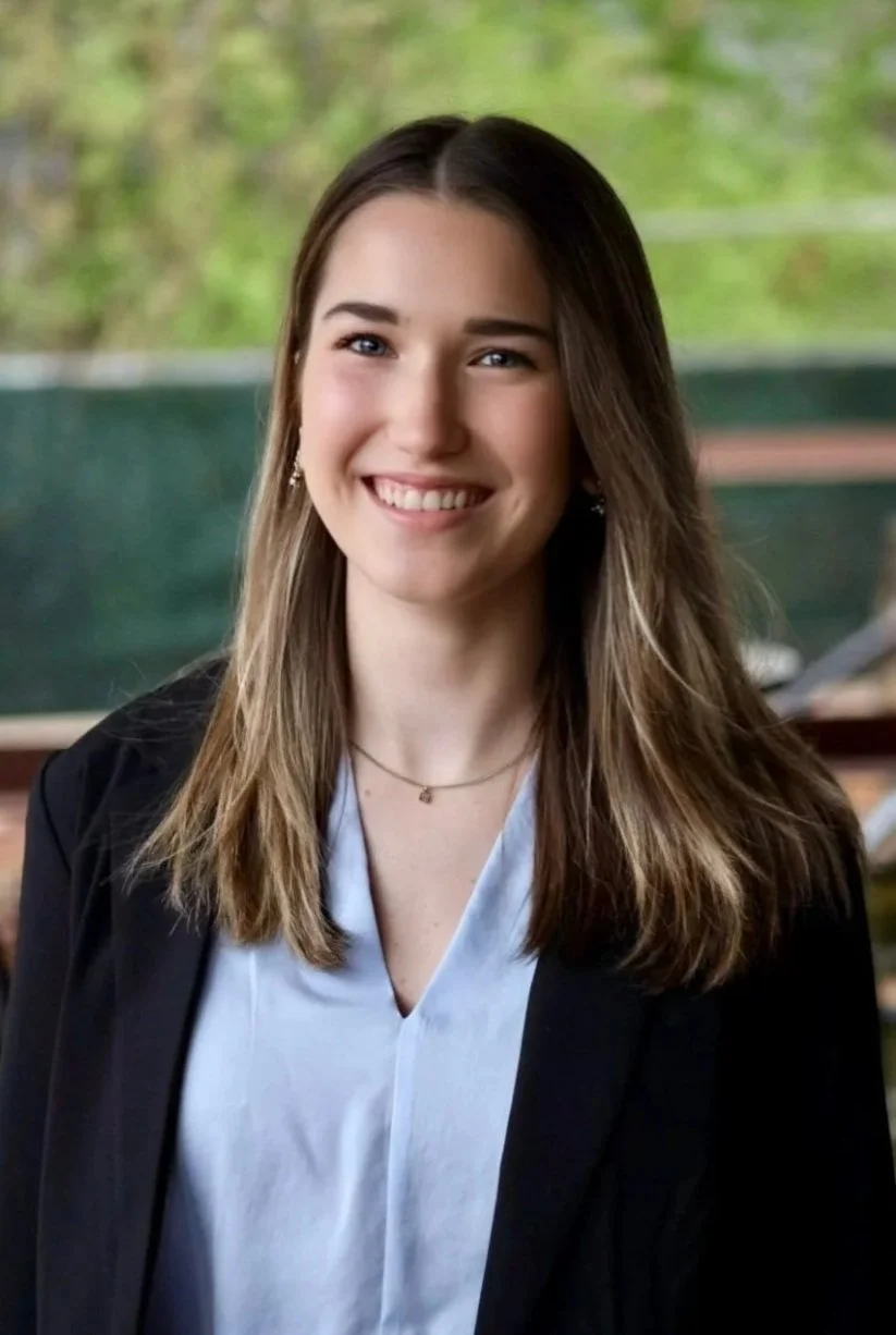 A young woman with long brown hair and blue eyes smiling, wearing a black blazer and light blue blouse, standing outdoors with green trees in the background.